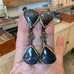 May include: A pair of dark blue gemstone earrings with gold-colored trim. The earrings have a geometric shape at the top and a teardrop shape at the bottom, connected by a floral design. The earrings are designed to dangle.