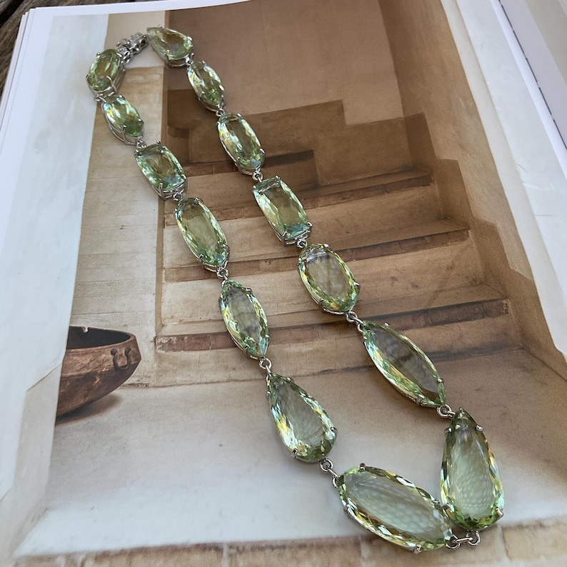 May include: A long silver chain necklace with large, faceted, light green gemstone pendants. The necklace is displayed on a textured surface.