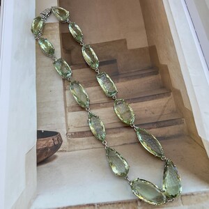May include: A long necklace with a silver chain and large, faceted, light green gemstones. The necklace is laid out on a brown and white patterned surface.