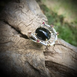 Peut inclure: Une bague en argent avec une pierre pr&eacute;cieuse noire sertie dans un design en forme de cage. La bague est orn&eacute;e de petites pierres pr&eacute;cieuses roses.