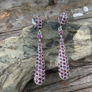May include: A pair of silver drop earrings with a cluster of small, round, deep red gemstones. The earrings have a teardrop shape, with a smaller cluster of gemstones at the top and a single gemstone in the middle.