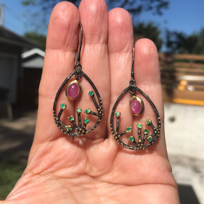 May include: A pair of dangle earrings with a pink gemstone and green gemstones set in a black and gold metal frame. The earrings are shaped like a teardrop with a delicate branch design.