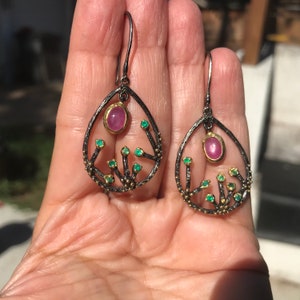May include: A pair of dangle earrings with a delicate, tree-like design. The earrings are made of black metal with gold accents and feature pink and green gemstones.