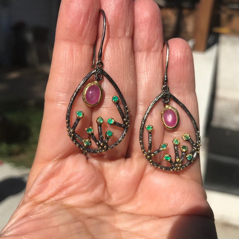 May include: A pair of dangle earrings with a delicate, tree-like design. The earrings are made of black metal with gold accents and feature pink and green gemstones.