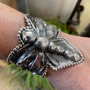 May include: A silver cuff bracelet with a floral design. The bracelet features a large, textured flower with detailed petals and a beaded border. The cuff is worn on a wrist.