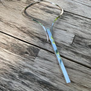 May include: A silver-toned necklace with a geometric design. The necklace features a curved upper portion and a long, rectangular pendant. The metal reflects the surrounding environment. The necklace is displayed on a wooden surface.