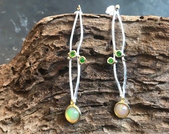 May include: A pair of silver and gold earrings with a white opal gemstone and green gemstones. The earrings are long and dangle.