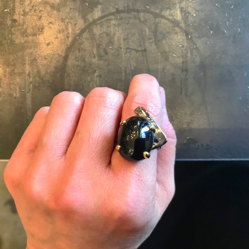 May include: A black gemstone ring with gold accents. The ring is set in a geometric design with a large oval-shaped stone.