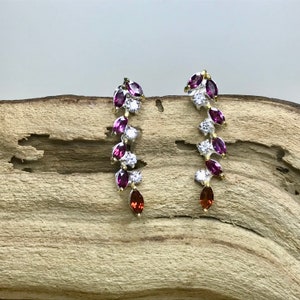 Puede incluir: Un par de pendientes colgantes de plata con una cascada de piedras preciosas p&uacute;rpuras y rojas y brillantes transparentes.