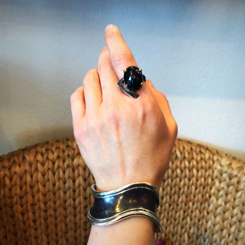 May include: A silver cuff bracelet with a wavy design. The bracelet is worn on a person's wrist. The person is also wearing a black ring with a gold setting.