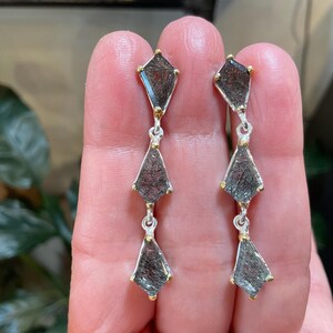 K&ouml;nnte beinhalten: Ein Paar silberne Ohrringe mit jeweils drei schwarzen und goldenen Rutilquarzsteinen. Die Steine sind facettiert und haben eine geometrische Form.