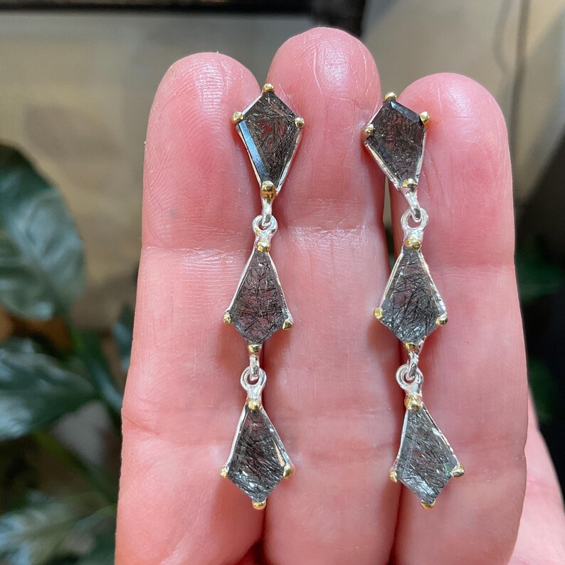 K&ouml;nnte beinhalten: Ein Paar silberne Ohrringe mit jeweils drei schwarzen und goldenen Rutilquarzsteinen. Die Steine sind facettiert und haben eine geometrische Form.