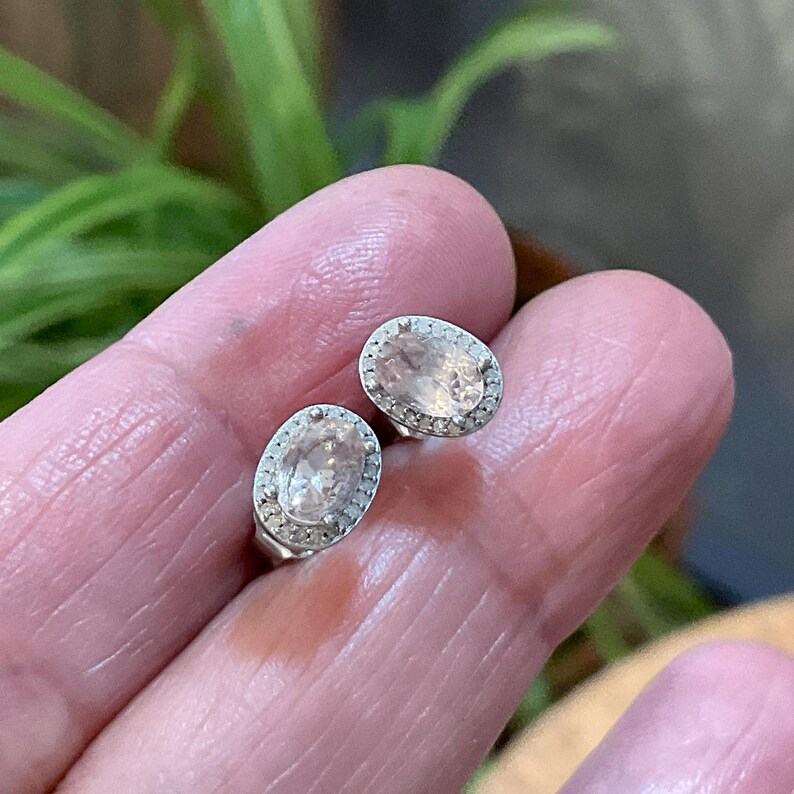 May include: A pair of oval-shaped stud earrings. Each earring features a light peach-colored gemstone surrounded by a halo of small, clear stones. The earrings are set in a silver-colored metal.