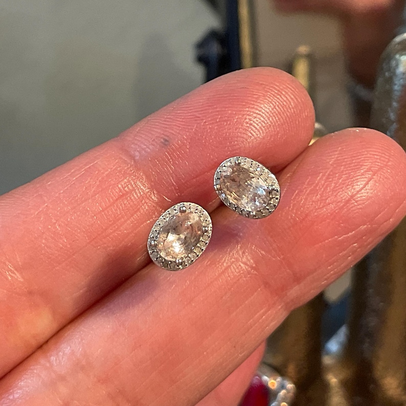 May include: A pair of oval-shaped stud earrings. Each earring features a light peach-colored center stone surrounded by a halo of small, clear stones. The earrings are set in a silver-colored metal.