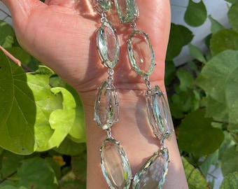 May include: A silver chain necklace with large, faceted, light green gemstone pendants. The necklace is on a person's wrist.