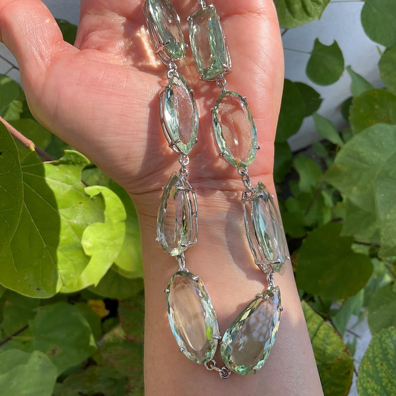 May include: A silver chain necklace with large, faceted, light green gemstone pendants. The necklace is on a person's wrist.