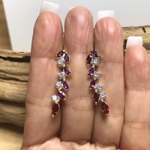 Puede incluir: Un par de pendientes colgantes con acabado plateado y dorado. Los pendientes presentan un dise&ntilde;o en cascada de peque&ntilde;as piedras de color p&uacute;rpura y transparentes en forma de pera.