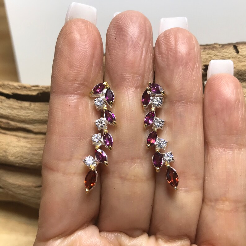 Puede incluir: Un par de pendientes colgantes con acabado plateado y dorado. Los pendientes presentan un dise&ntilde;o en cascada de peque&ntilde;as piedras de color p&uacute;rpura y transparentes en forma de pera.