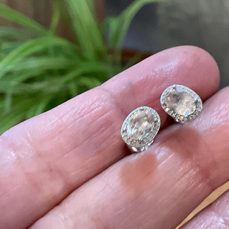 May include: A pair of oval-shaped stud earrings. Each earring features a clear, oval-cut gemstone surrounded by a halo of smaller, clear stones. The earrings are set in a silver-colored metal, creating a sparkling and elegant appearance.