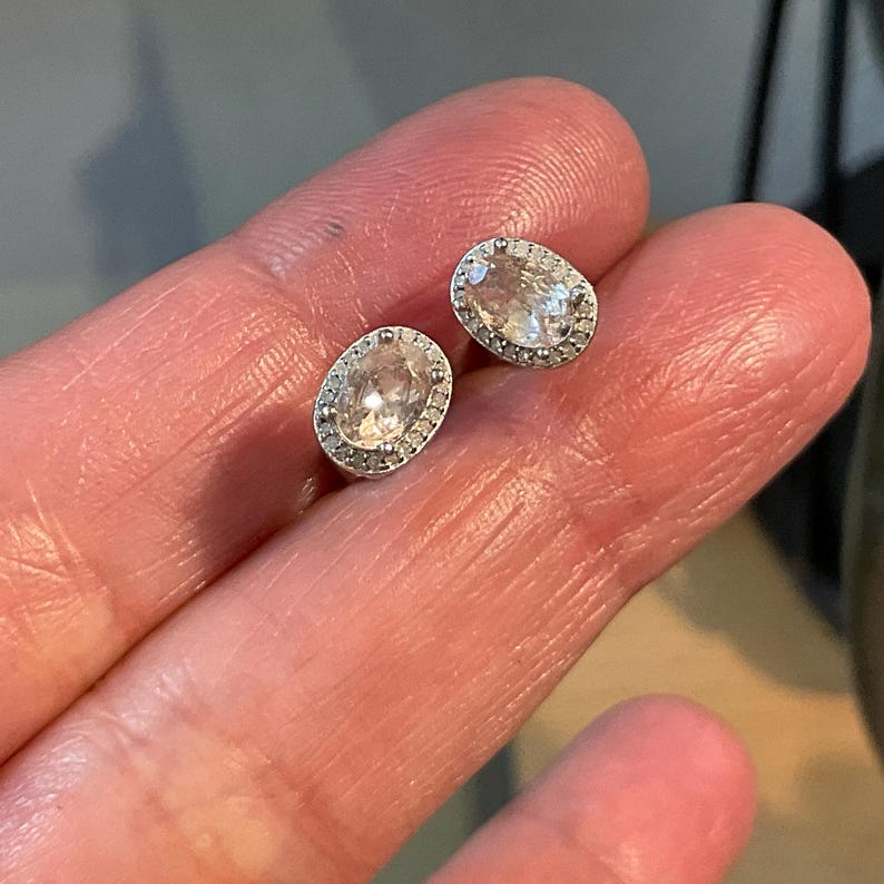 May include: Two silver-toned oval stud earrings. Each earring features a clear, oval-shaped stone surrounded by a halo of smaller, clear stones. The earrings are displayed on a light-toned background.