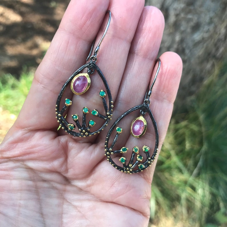 May include: A pair of dangle earrings with a black oxidized silver setting and a pink oval gemstone. The earrings feature a delicate design of branches with small green gemstones.