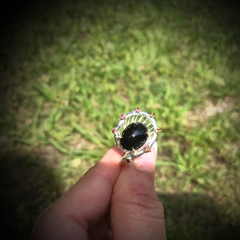 Peut inclure: Une bague en argent avec une pierre ovale noire entour&eacute;e de petites pierres roses.