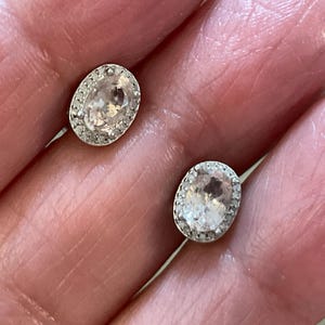 May include: A pair of oval-shaped stud earrings. Each earring features a light pink gemstone surrounded by a halo of small, clear stones. The earrings are set in a silver-colored metal.