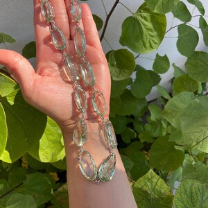 May include: A long necklace made of faceted, light green gemstone beads. The beads are oval shaped and are strung on a silver chain.