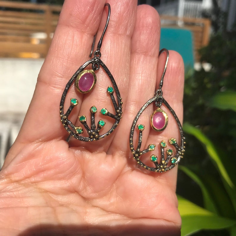 May include: A pair of dangle earrings with a teardrop-shaped design. The earrings are made of black metal with gold accents and feature a pink gemstone and several smaller green gemstones.