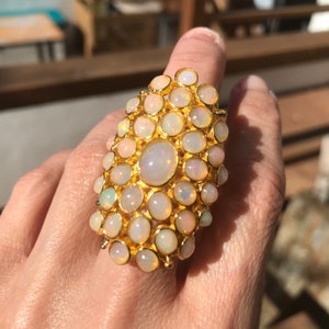 May include: A gold ring featuring a cluster of white opals. The ring is oval shaped and has a large central opal surrounded by smaller opals.
