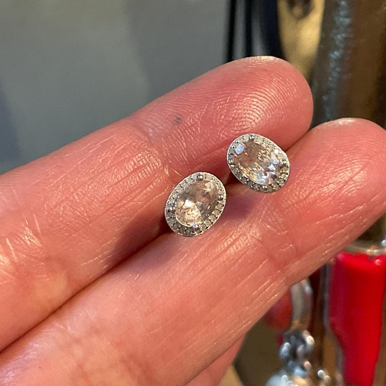 May include: A pair of oval-shaped stud earrings. Each earring features a light peach-colored gemstone surrounded by a halo of small, clear stones. The earrings are set in a silver-colored metal.