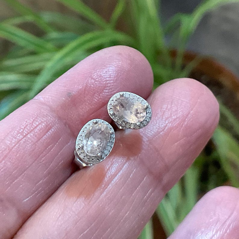 May include: A pair of oval-shaped stud earrings. Each earring features a light peach-colored gemstone surrounded by a halo of small, clear stones. The earrings are set in a silver-colored metal.