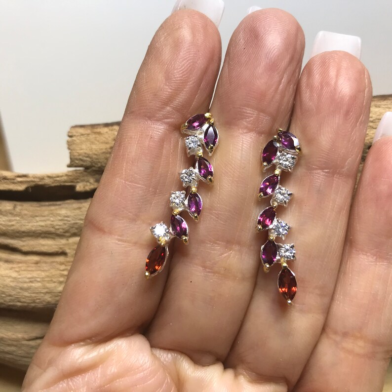Puede incluir: Un par de pendientes colgantes con una cascada de piedras preciosas p&uacute;rpuras y rojas y piedras de circonita c&uacute;bica transparentes. Los pendientes est&aacute;n engastados en plata con detalles dorados.