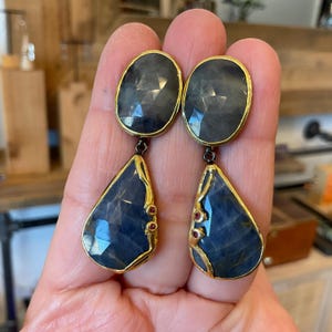 May include: A pair of handcrafted earrings featuring large, faceted, oval-shaped gray gemstones at the top, connected to teardrop-shaped blue gemstones. The earrings have gold-colored trim and small red accents, showcasing a unique jewelry design.