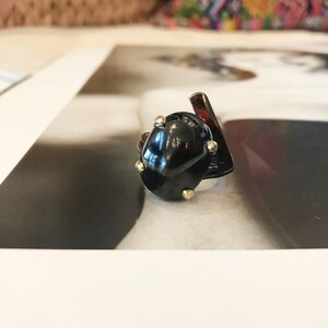 May include: A black ring with a large, irregularly shaped black gemstone. The ring is accented with small gold studs around the gemstone.