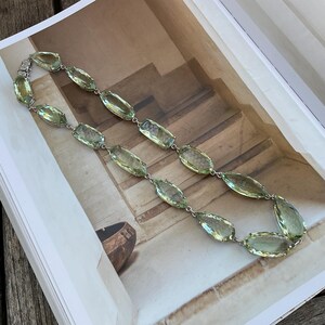 May include: A long necklace made of light green faceted stones connected by silver chain links. The necklace is laid out on a book with a brown wooden surface in the background.