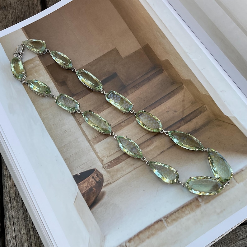 May include: A long necklace made of light green faceted stones connected by silver chain links. The necklace is laid out on a book with a brown wooden surface in the background.