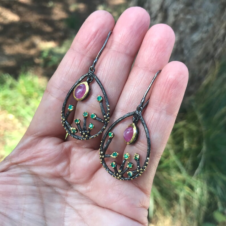 May include: A pair of dangle earrings with a black oxidized silver setting and gold accents. The earrings feature a pink gemstone and green gemstones set in a tree branch design.