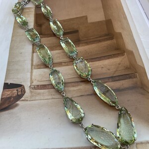 May include: A long silver chain necklace with large, faceted, light green gemstone drops. The necklace is made of silver and has a simple design.