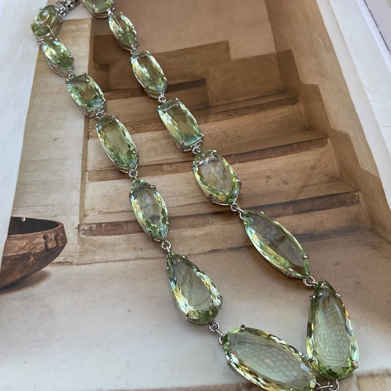 May include: A long silver chain necklace with large, faceted, light green gemstone drops. The necklace is made of silver and has a simple design.