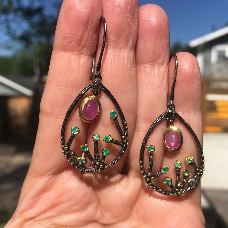 May include: A pair of dangle earrings with a black oxidized silver setting and gold accents. The earrings feature a pink gemstone and green gemstones set in a delicate branch design.