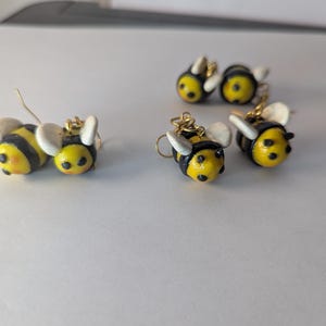 May include: Handmade bee earrings. These earrings feature a yellow body with black stripes, white wings, and gold-colored earring hooks. The earrings are small and detailed, with a cute, cartoonish design.