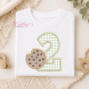 May include: A white t-shirt featuring an embroidered design of the number two in a light green and white gingham pattern, accompanied by a brown chocolate chip cookie with a bite taken out of it, positioned on the left side of the number.