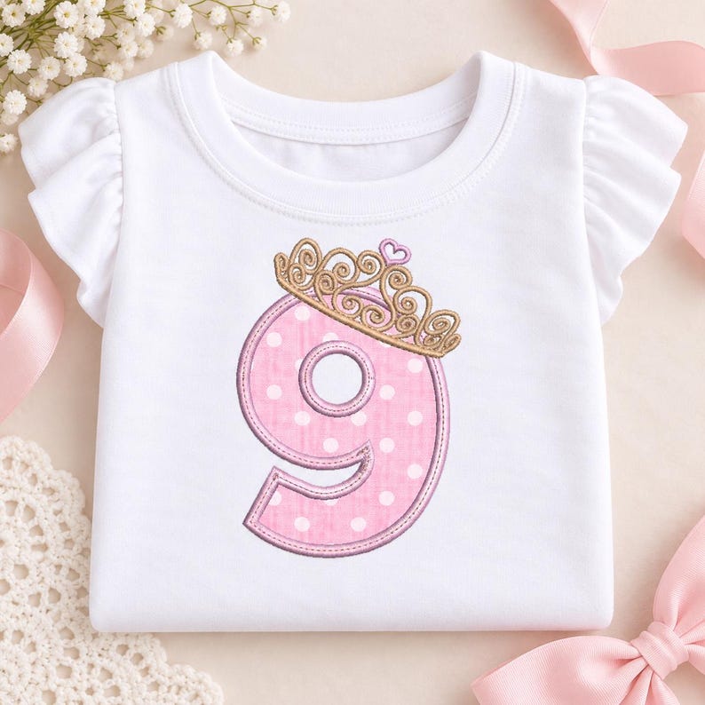 May include: A white short-sleeved shirt with ruffled sleeves featuring an embroidered pink number nine with white polka dots. The number is topped with a gold-colored crown design and a small pink heart accent, centered on the front.