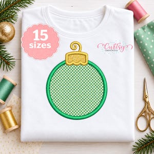 May include: A white t-shirt featuring a centered machine embroidery design of a round Christmas ornament. The ornament has a green gingham pattern center, a thick green border, and a textured gold cap. Text reads 15 sizes and Curtsy Embroidery.