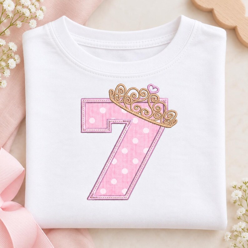 May include: A white short-sleeved t-shirt featuring a large embroidered number seven in pink with white polka dots. The number is topped with a gold-colored crown design that includes a small pink heart detail at the very top center.