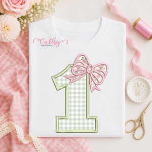 May include: A white t-shirt featuring an embroidered number one with a light green and white gingham pattern, outlined in light green thread. A pink and white floral patterned bow is attached to the top right of the number. The text Curtsy Embroidery is printed above the number.