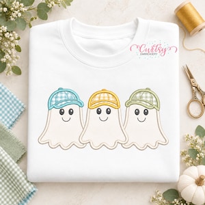 Peut inclure: Un sweat-shirt blanc à col rond orné d'une broderie représentant trois fantômes côte à côte. Chaque fantôme porte une casquette de baseball à carreaux vichy de couleur différente : bleu clair, jaune et vert sauge. Le texte Curtsy Embroidery est visible.