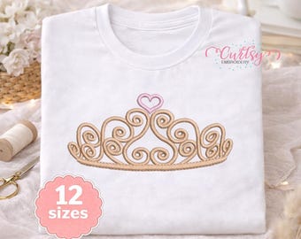 May include: A white t-shirt featuring an embroidered gold tiara design with a small pink heart at the top center. The shirt includes the text Curtsy EMBROIDERY in the upper right corner and a pink scalloped label stating 12 sizes.