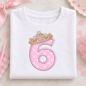 May include: A white short-sleeved t-shirt featuring an embroidered design of the number six in pink with white polka dots. The number is topped with a gold-colored crown with a small heart detail at the peak, outlined in dark pink thread.